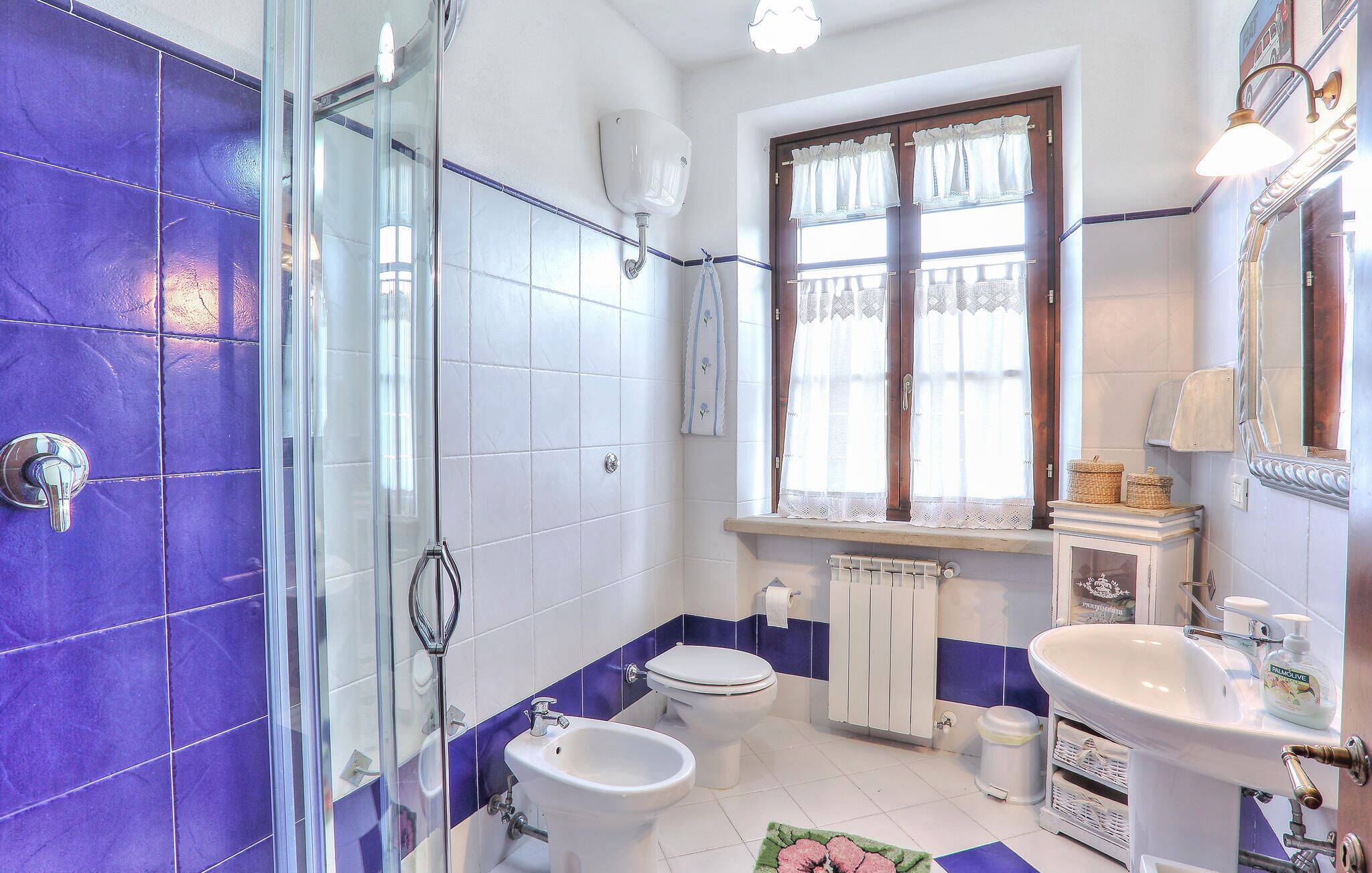 Casa de banho com azulejos roxos, cabine de duche em vidro, sanita, bidé, lavatório e janela com cortinas de renda.