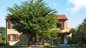 Une maison à deux étages, avec un grand arbre devant, un mur en pierre et une terrasse aménagée avec du mobilier d’extérieur.