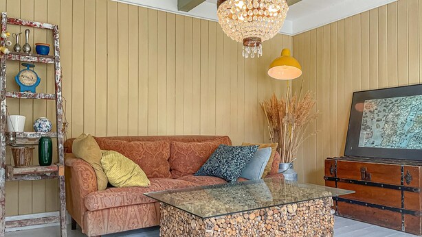 A living room with a patterned sofa, a glass coffee table, a wooden cabinet, a chandelier, and a wall-mounted shelf with decorative items. - 4 bedroom pet friendly home in Farsund (Farsund)
