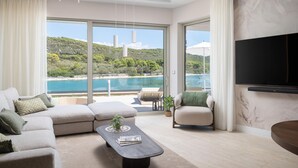 A modern living room with a sofa, armchair, and coffee table. Large windows offer a view of a beach and hills. - Stunning home in Kali (Kali)