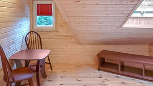 Une petite pièce mansardée aux murs en bois et au plafond en pente. On y trouve une fenêtre avec des stores rouges, un banc en bois et deux chaises.