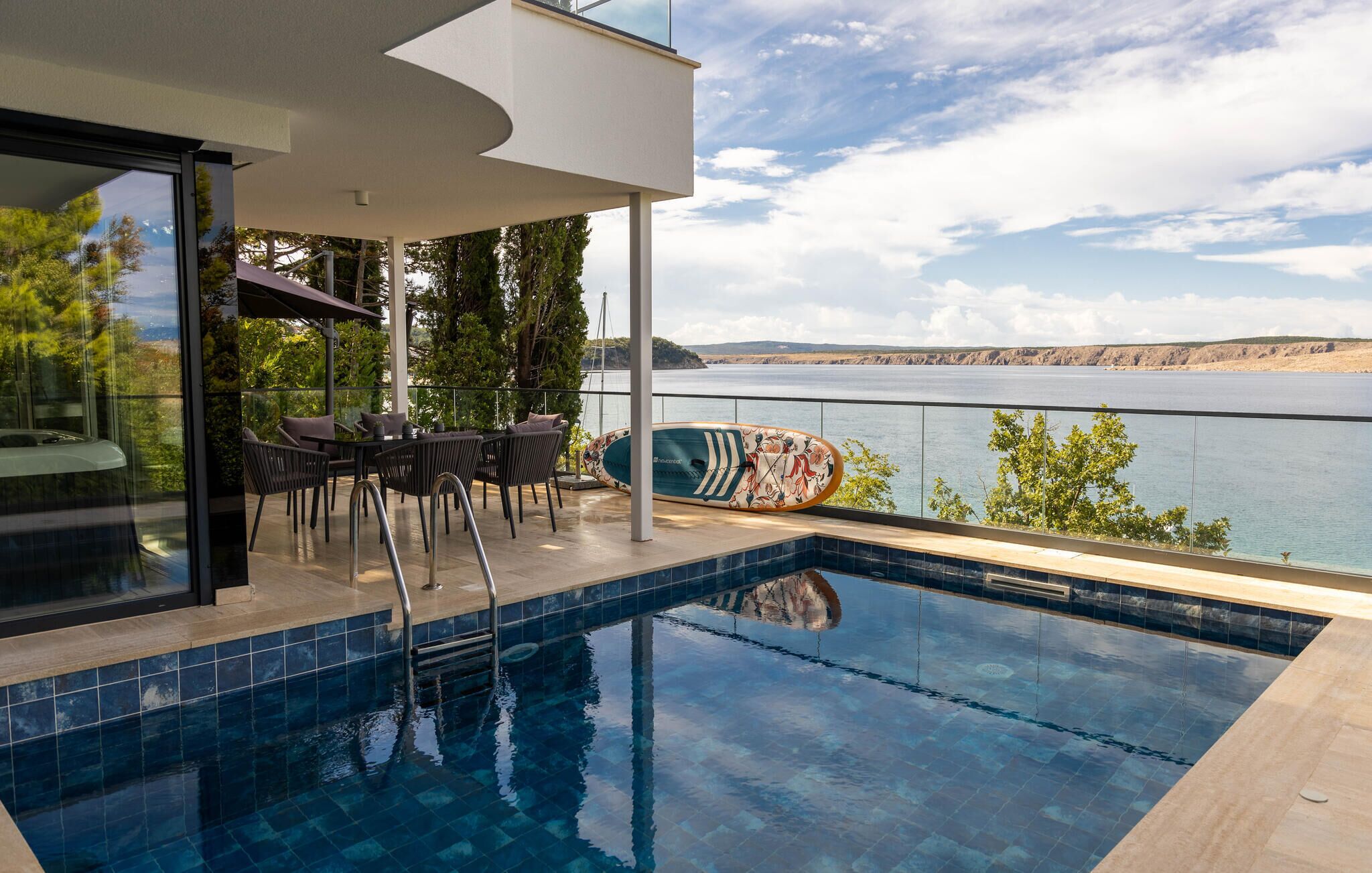 Uma área moderna de piscina ao ar livre com vista para um lago e colinas, apresentando um espaço de descanso com cadeiras e uma mesa, e um prancha de surf encostada na grade.