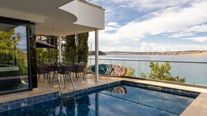 Uma área moderna de piscina ao ar livre com vista para um lago e colinas, apresentando um espaço de descanso com cadeiras e uma mesa, e um prancha de surf encostada na grade.