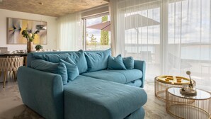 Uma sala moderna com um sofá azul, uma área de jantar com mesa e cadeiras, e janelas amplas com vista para o exterior.