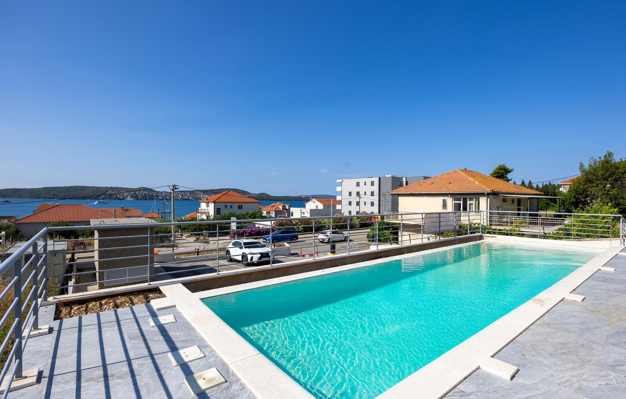 Una piscina amb aigua cristal·lina blava, envoltada d'una tanca metàl·lica, amb vista al mar i edificis al fons.