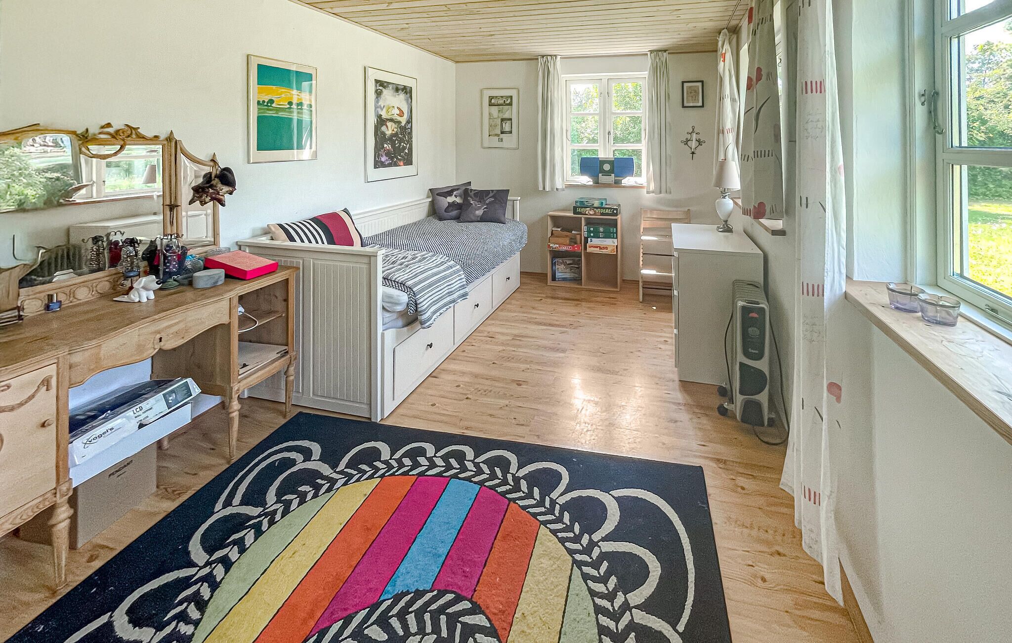 Une pièce compacte et bien éclairée, comprenant un lit, un bureau et une petite kitchenette. Les murs sont ornés de tableaux encadrés et il y a une fenêtre avec des rideaux.