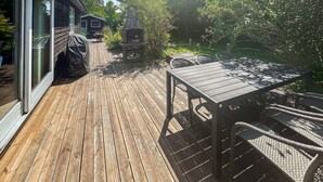 Una terraza de madera con una mesa y sillas, rodeada de follaje y una estructura pequeña al fondo.