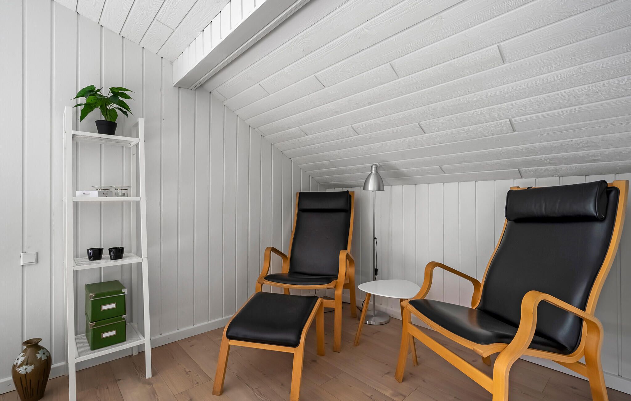 Ein Zimmer mit zwei schwarzen Ledersesseln, einem kleinen weißen Tisch, einer Stehlampe, einer weißen Regaleinheit mit grünen Boxen und einer Topfpflanze.