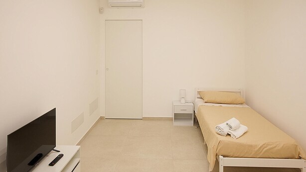 Una stanza piccola e pulita con un letto, un comodino, una TV e un condizionatore d'aria.