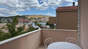Un balcon avec une table et des chaises, donnant sur une ville côtière avec des maisons à étages et une colline en arrière-plan.