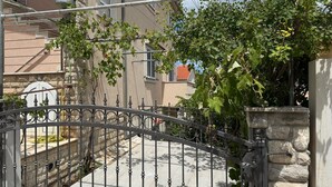 Une porte en fer forgé, un mur en pierre et, au fond, un bâtiment avec un toit recouvert de tuiles.