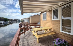 Une terrasse en bois avec des sièges extérieurs, donnant sur un canal, et bordée d’une rangée de bâtiments similaires.