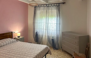 A bedroom with a bed, a dresser, a nightstand with a lamp, and a window with a curtain.