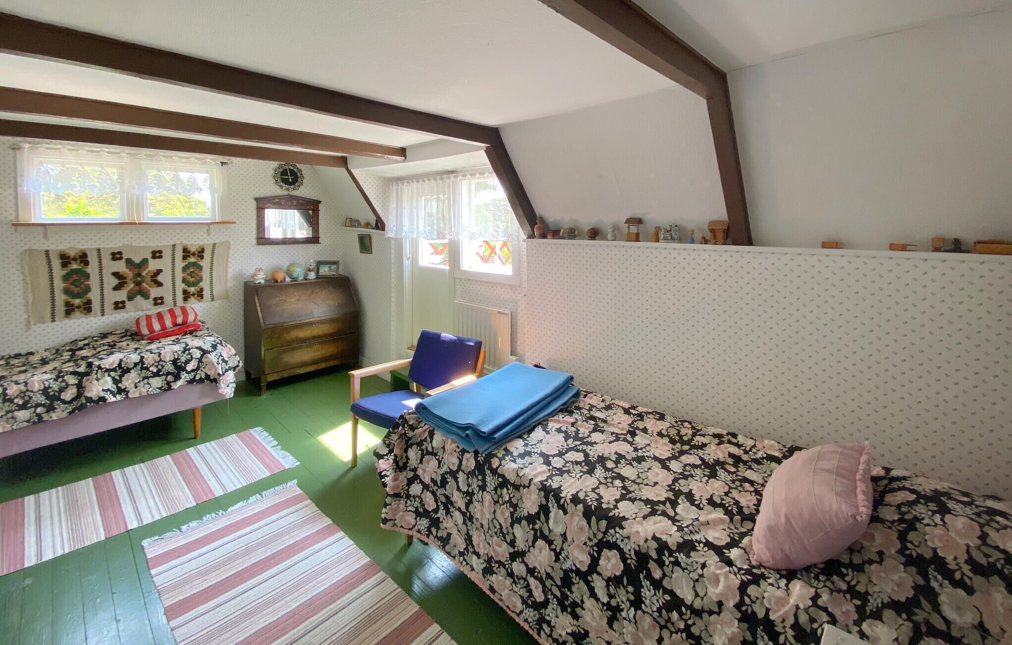 A cozy room with two single beds, a wooden dresser, a blue chair, and a patterned rug.