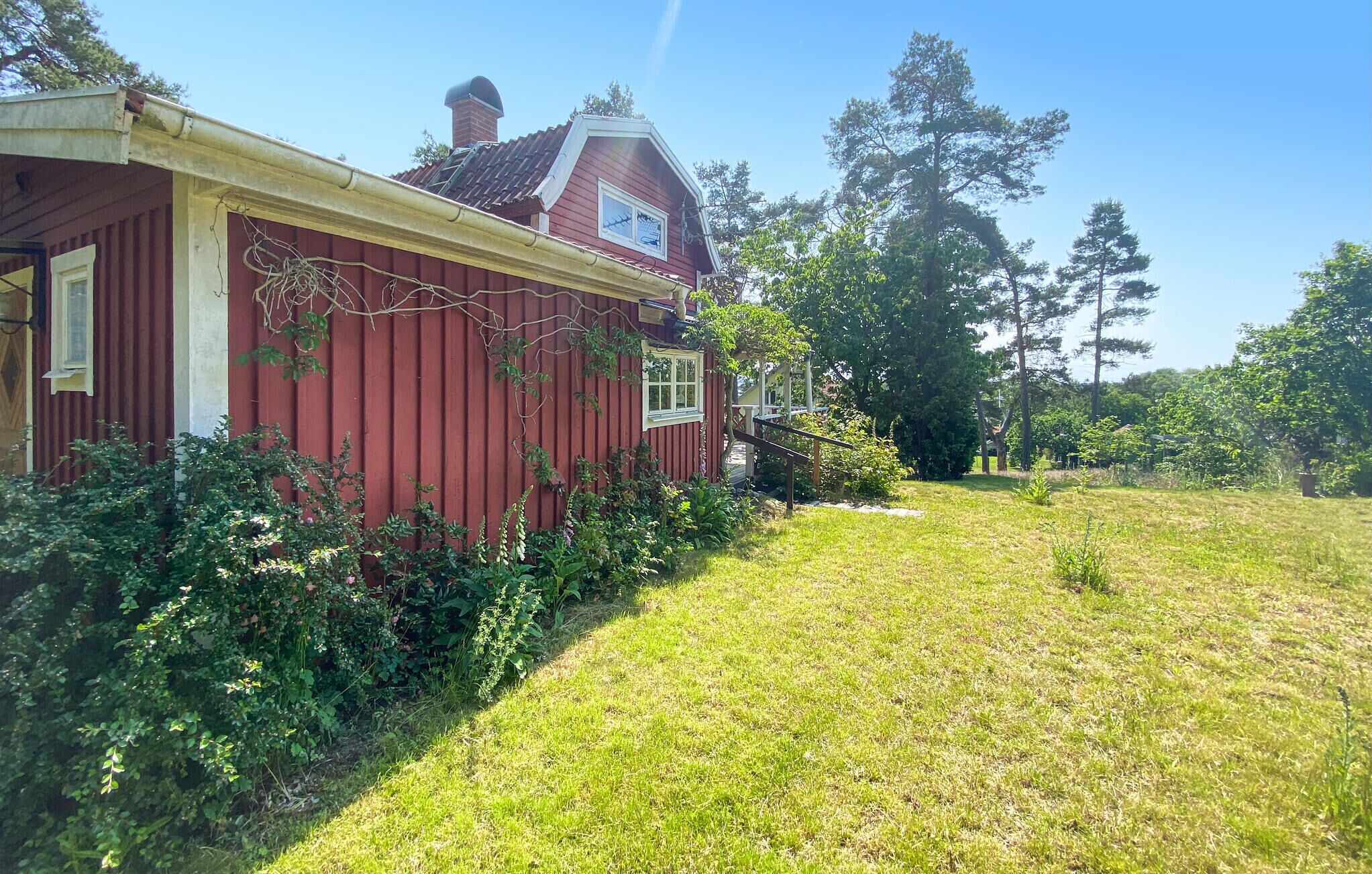 Une maison en bois rouge, avec un toit en pente et une cheminée, entourée d’une cour verdoyante et d’arbres.