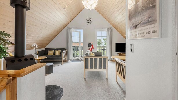 A cozy loft-style room with a high ceiling, wooden beams, a black stove, a sofa, a dining table, and a TV. - Stunning home in Gråsten with sauna (Gråsten)