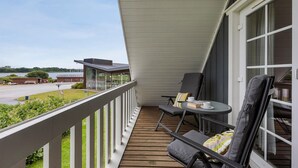 A balcony with a wooden floor, two chairs, a small table with a cup and a book, and a view of a building and a parking lot. - Stunning home in Gråsten with sauna (Gråsten)
