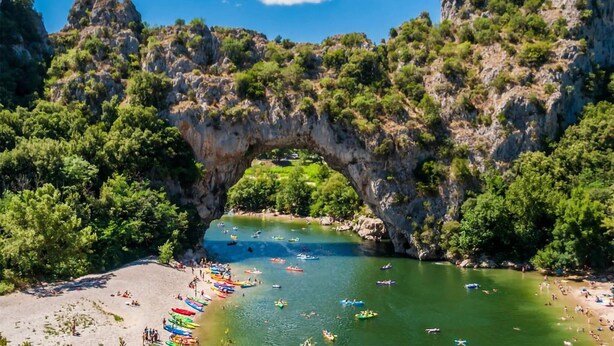 Une arche naturelle formée par la roche, surplombant une plage où l’on voit des kayaks et des personnes.