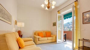 A living room with a sofa, a table, and a lamp. There is a door leading to a balcony with a view of buildings. - 2 bedroom amazing apartment in Recco (Recco)