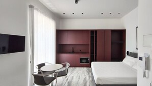 A modern hotel room with a bed, a desk, a chair, a TV, and a minibar.