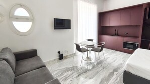 A modern hotel room with a grey sofa, a round dining table with chairs, a TV mounted on the wall, and a kitchen area with a microwave and oven.