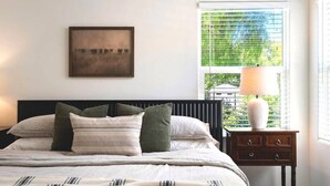 Une chambre à coucher avec un lit, des oreillers, une table de chevet avec une lampe, une fenêtre avec des stores et un tableau encadré au mur.