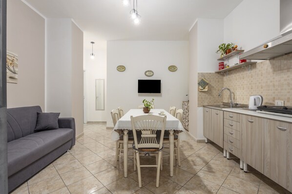 A compact living space with a dining table, chairs, a sofa, a kitchen area with cabinets and a sink, and a flat-screen TV mounted on the wall.