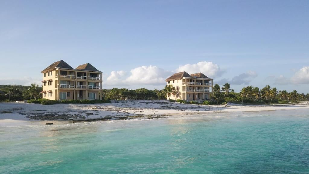 Dos edificios frente a la playa con balcones, una playa de arena y palmeras.