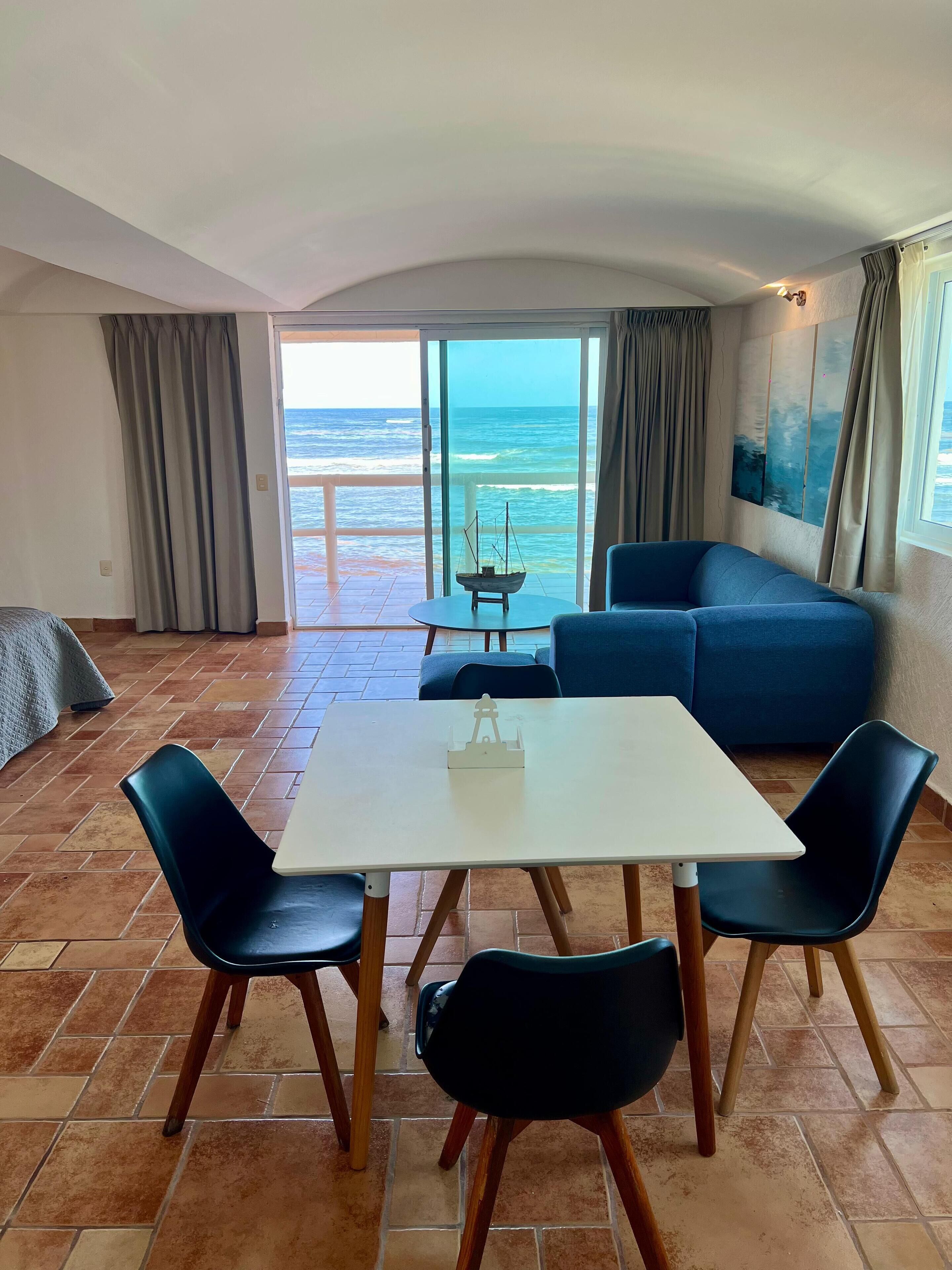 A room with a white table and four chairs, a blue sofa, and a view of the ocean through large glass doors.