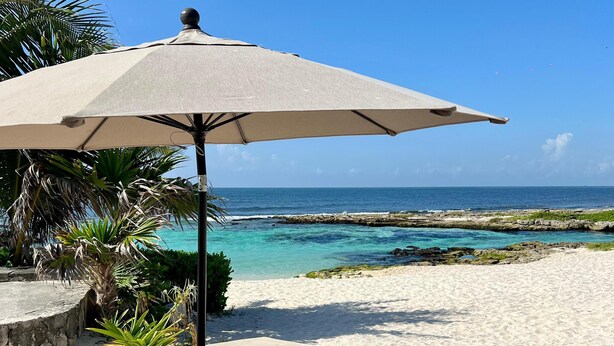 A beachside seating area with a table and an umbrella, overlooking a clear blue sea and rocky shoreline. - Caleta Tankah (Tulum)