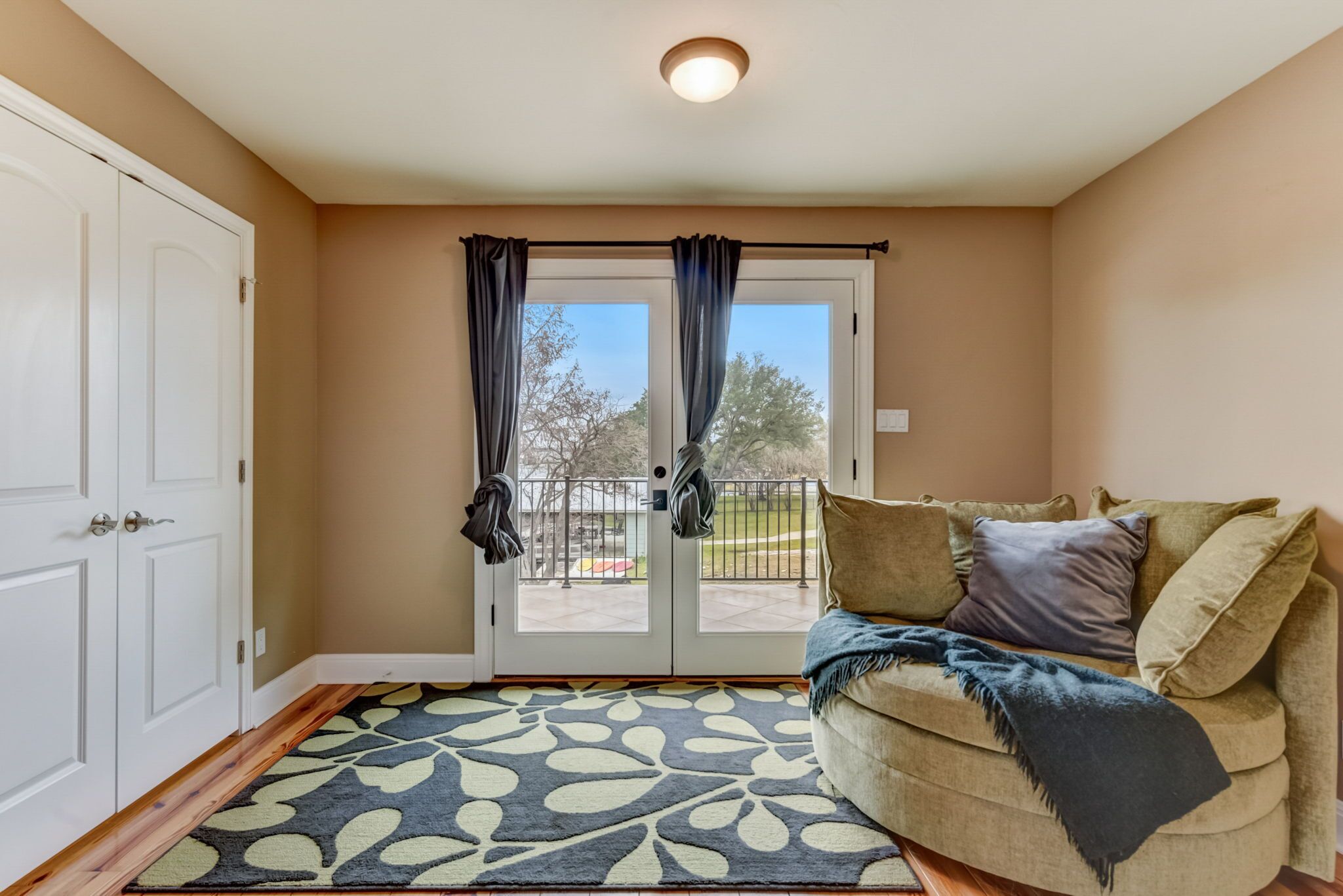 A room with a beige wall, a white door, a sliding glass door with curtains, a beige sofa with cushions, and a patterned rug.