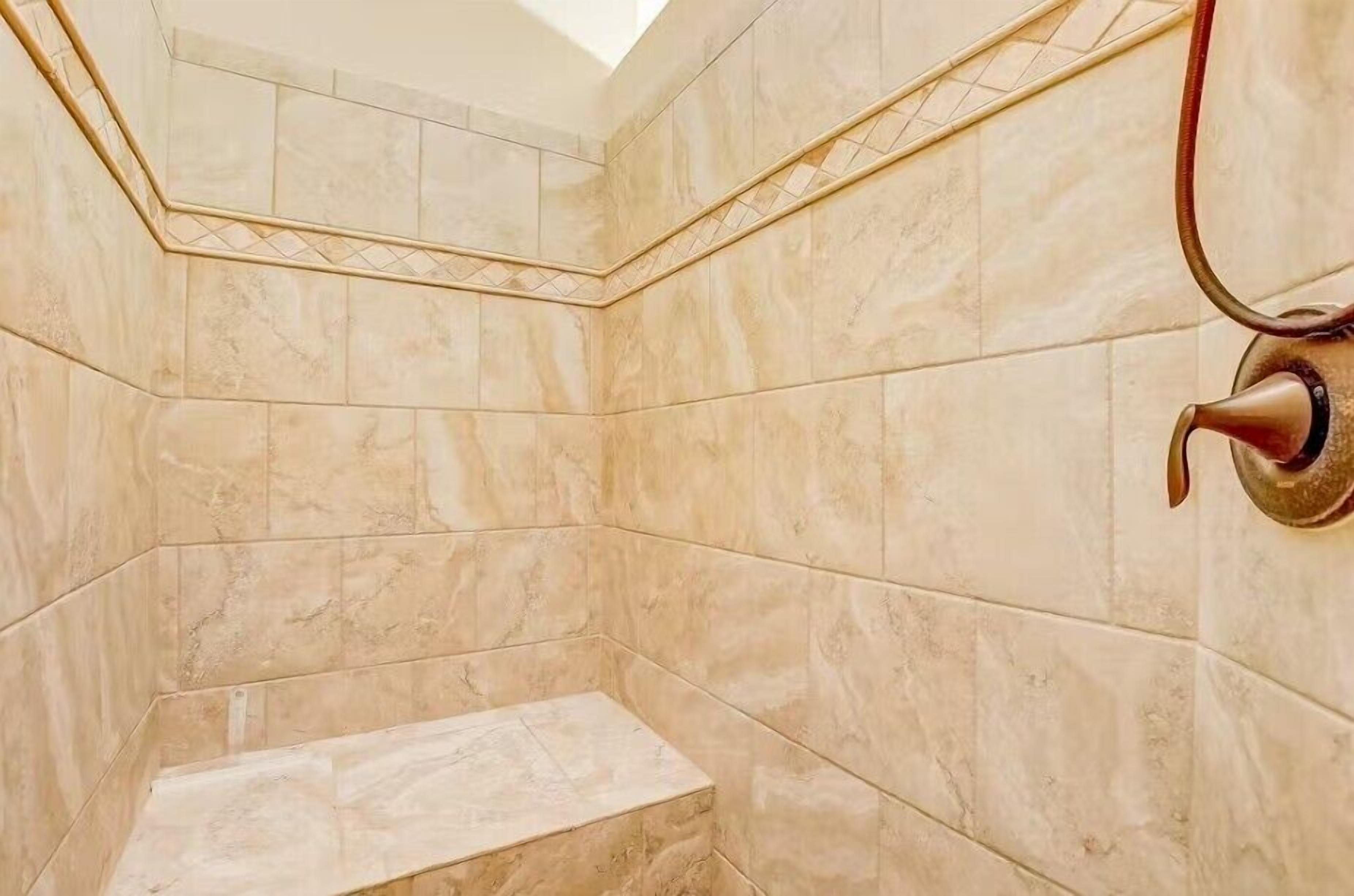 Un coin d’une salle de bain avec des carreaux de marbre beige, une pomme de douche et un banc.