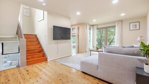 Un salón espacioso con una escalera de madera, un sofá gris y un televisor de pantalla plana montado en la pared.