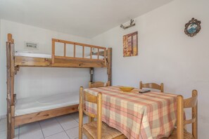 Uma cama de beliche com estrutura de madeira, uma mesa com toalha quadriculada e um quadro na parede.