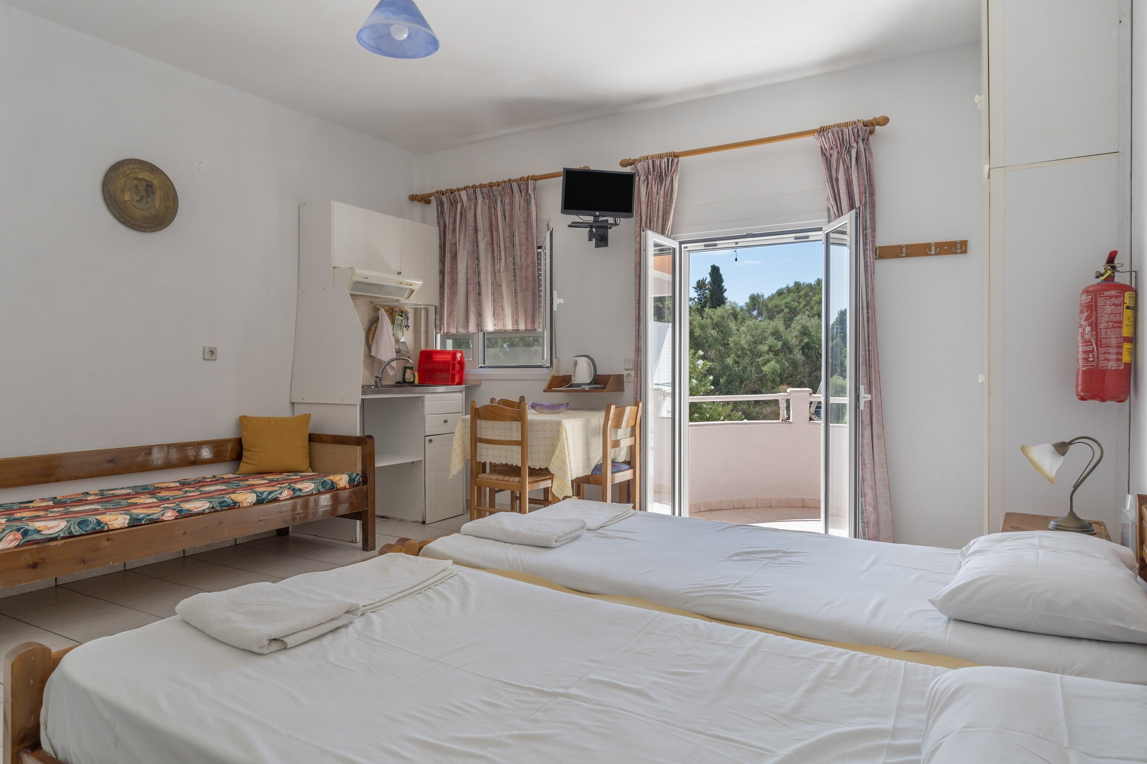 Een kamer met twee bedden, een bureau, een stoel, een televisie en een balkon met een brandblusser.