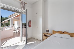 Un dormitorio con cama, una mesita de noche, una lámpara, un armario y un ventanal corredero que da a un balcón con mesa y sillas.