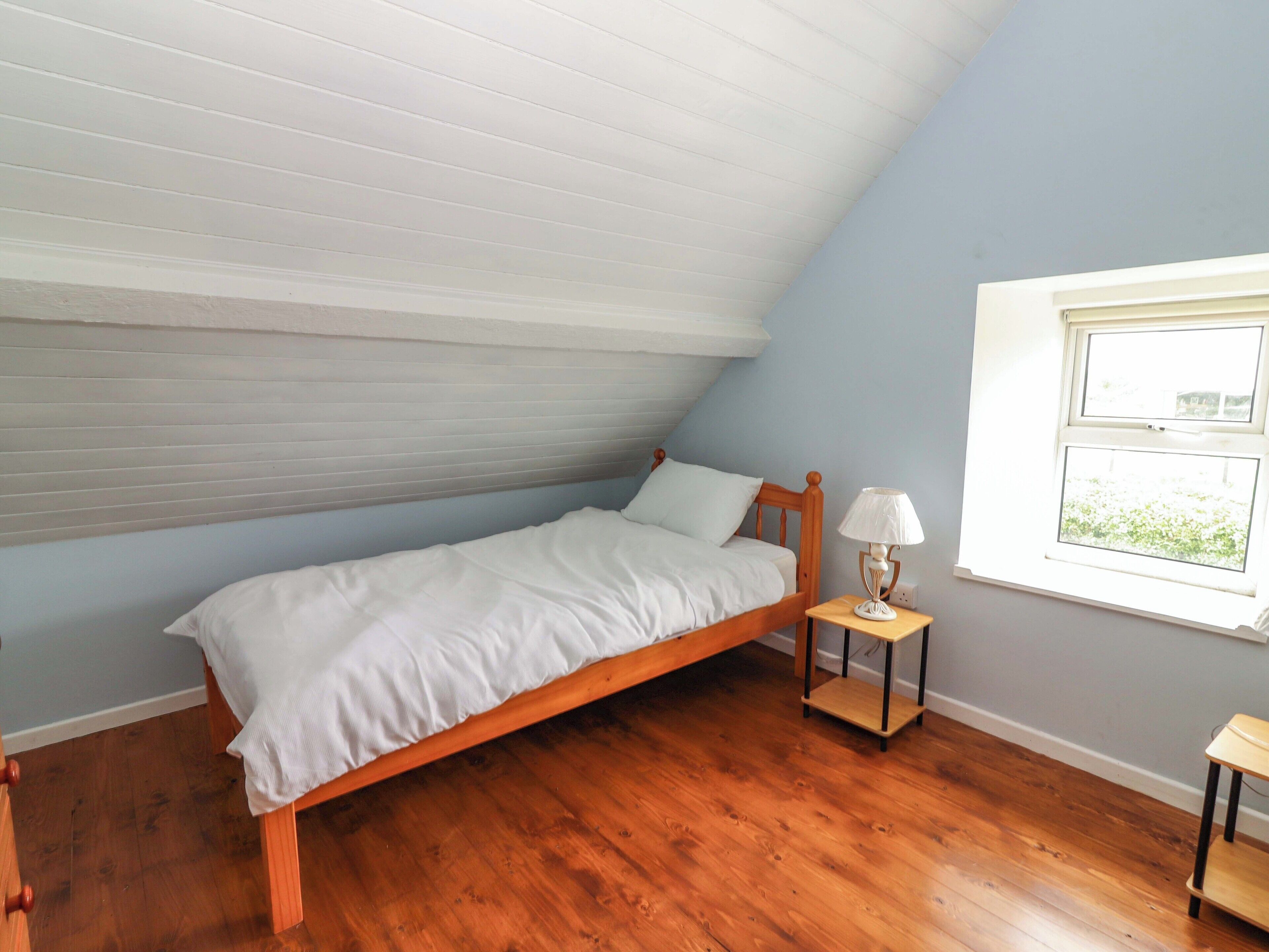 Une petite chambre avec un lit simple, un plafond en pente, un plancher en bois, une table de chevet avec une lampe et une fenêtre donnant sur un beau paysage.