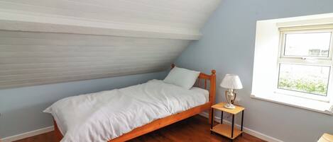 Une petite chambre avec un lit simple, un plafond en pente, un plancher en bois, une table de chevet avec une lampe et une fenêtre donnant sur un beau paysage.