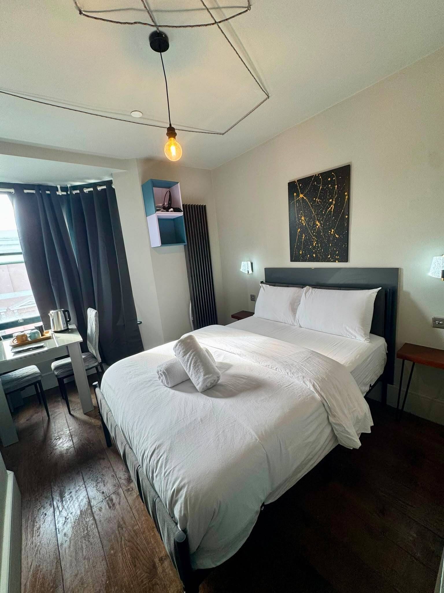 Una habitación de hotel con cama, escritorio, silla, radiador, ventana con cortinas y una lámpara de pared.