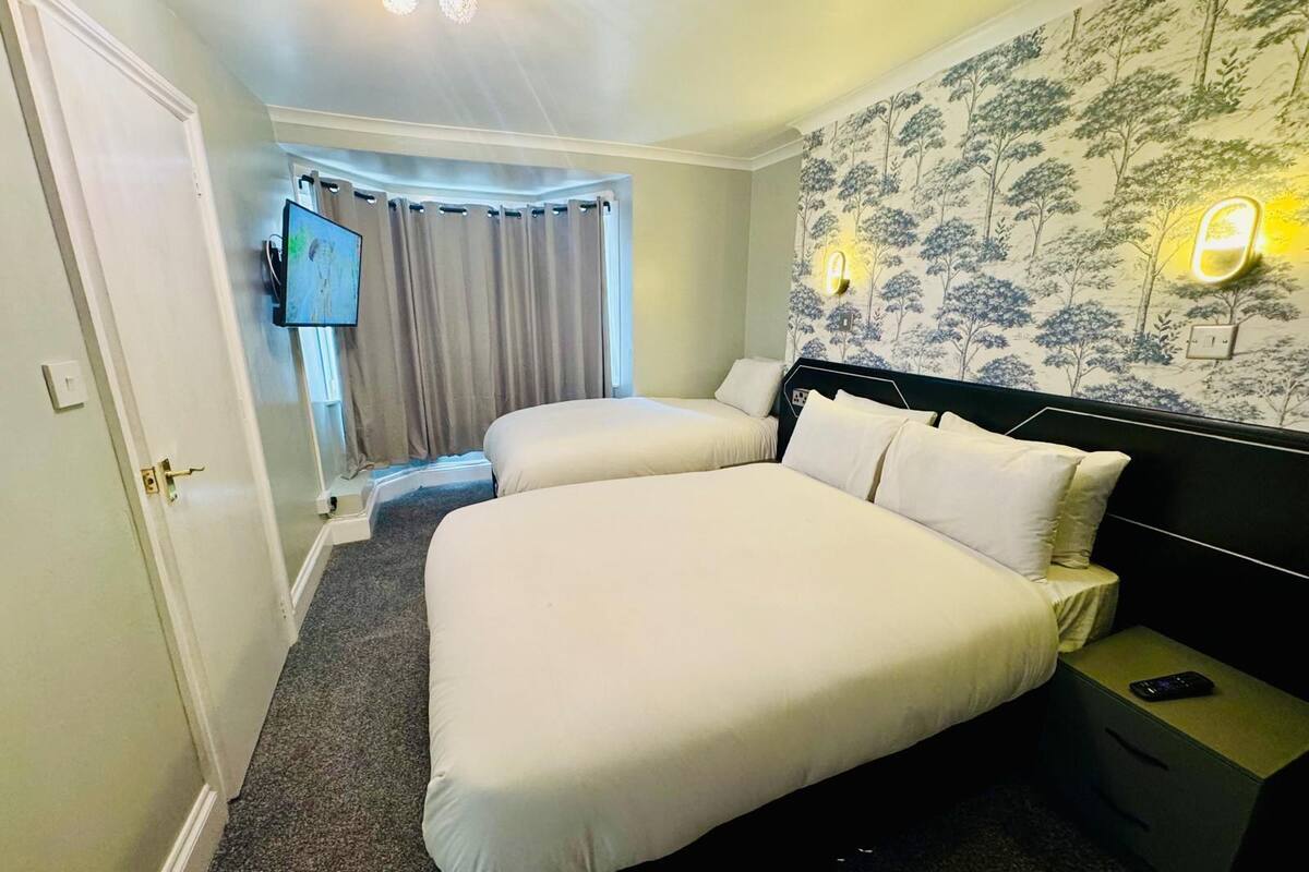 A hotel room with two beds, a television, a wall-mounted light, and a patterned wallpaper.