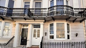 A multi-story building with a classic architectural style, featuring a prominent entrance with a door labeled "Sachi Hotel." The building has a symmetrical facade with decorative elements and a black wrought-iron balcony railing. - Sachi Hotel (Brighton)