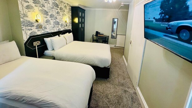 A hotel room with two beds, a desk, a chair, a television, and a wall with a floral wallpaper. - Sachi Hotel (Brighton)