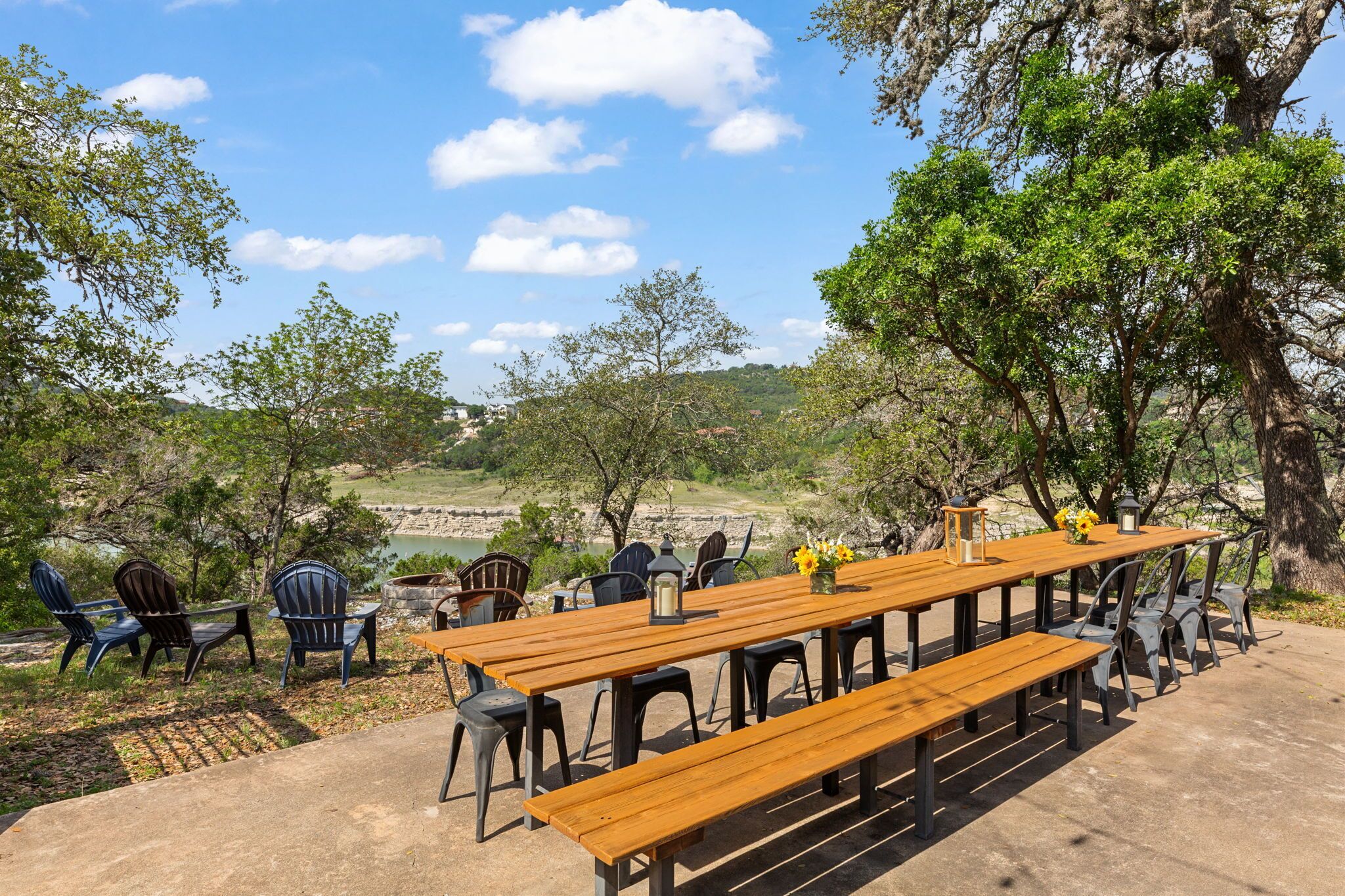 An outdoor dining area with a long wooden table and chairs, surrounded by trees and a view of a lake.