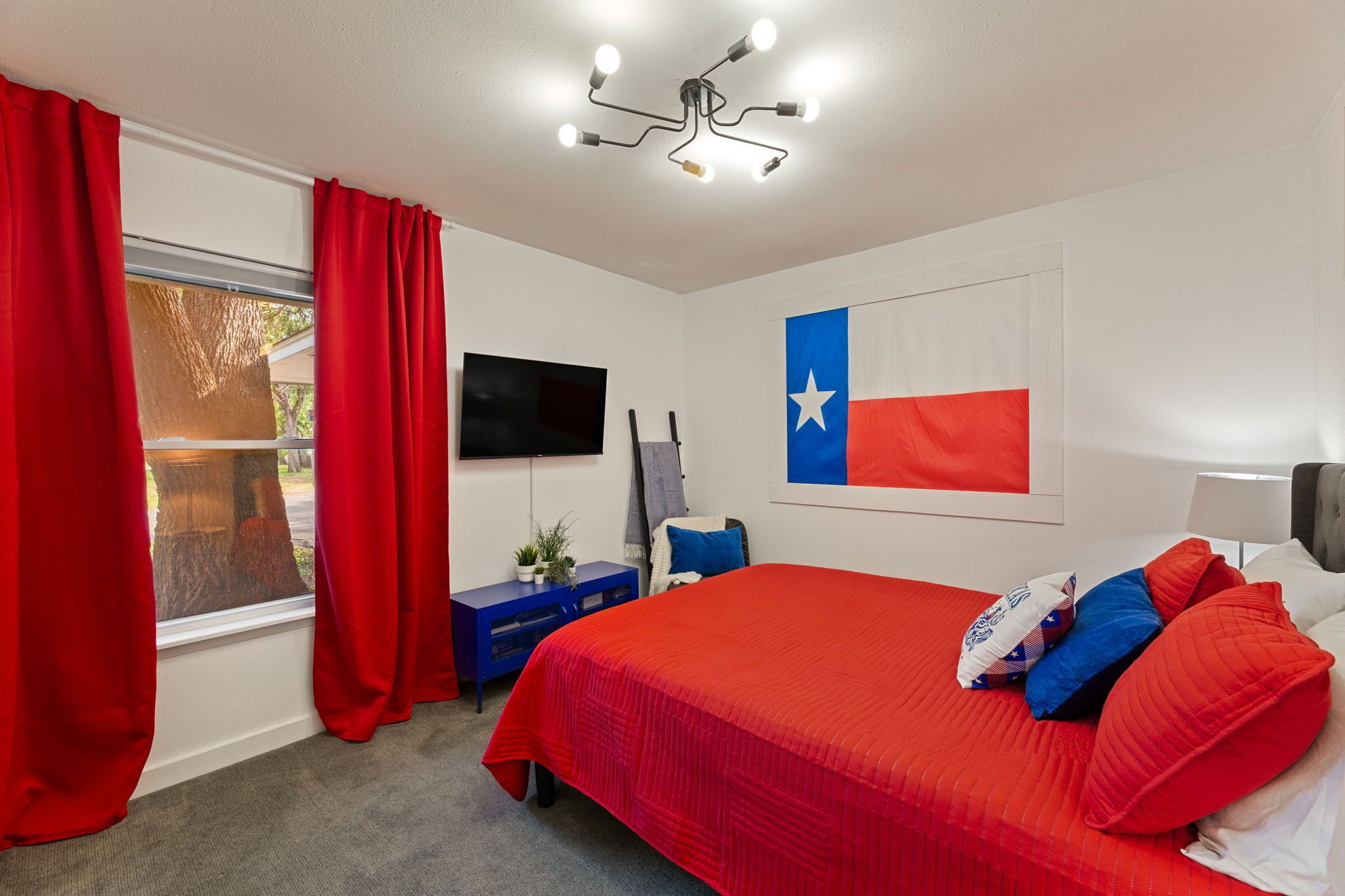 A hotel room with a large bed, red curtains, a Texas flag, a TV, and a desk with a lamp.