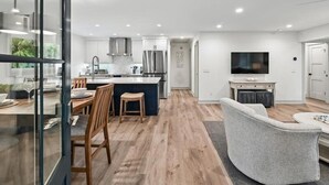 Een moderne keuken met een centraal eiland, een eethoek met een glazen scheiding, een woonkamer met een grijze bank en een flatscreen tv aan de muur bevestigd.