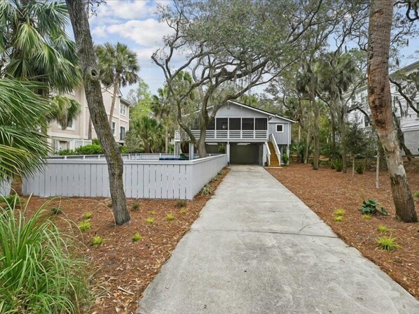 A residential property with a concrete driveway leading to a garage, surrounded by mature trees and a white fence.