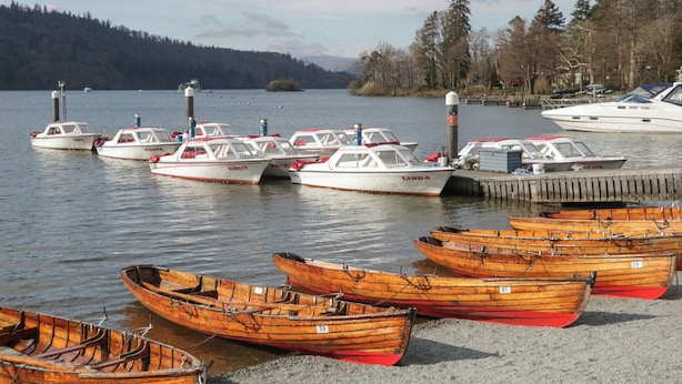 Um cais à beira do lago com vários barcos, incluindo barcos a motor e barcos a remo, ancorados perto da margem.