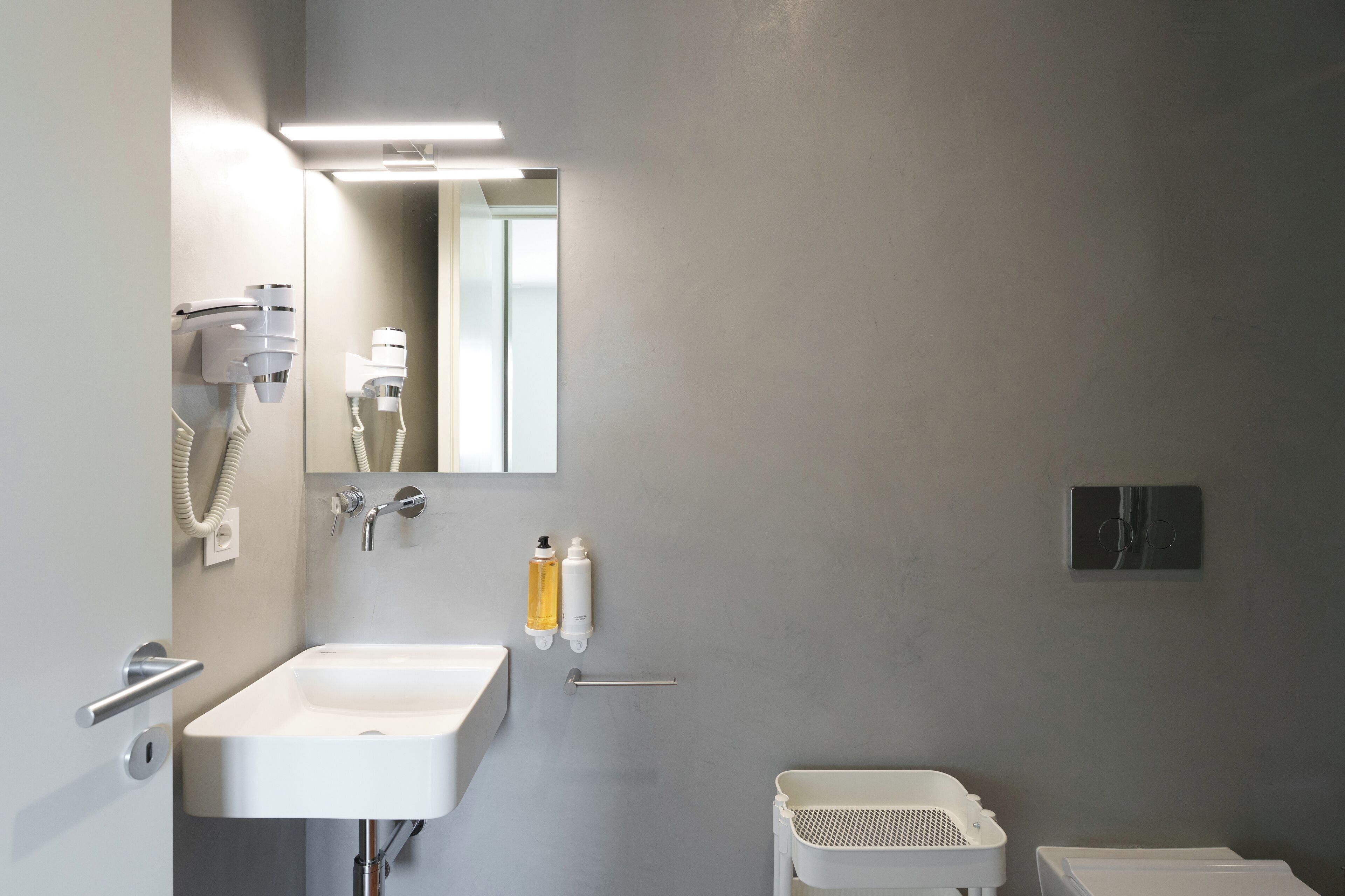 Een moderne badkamer met een witte wastafel, een spiegel, een haardroger en een klein wit wasmandje.