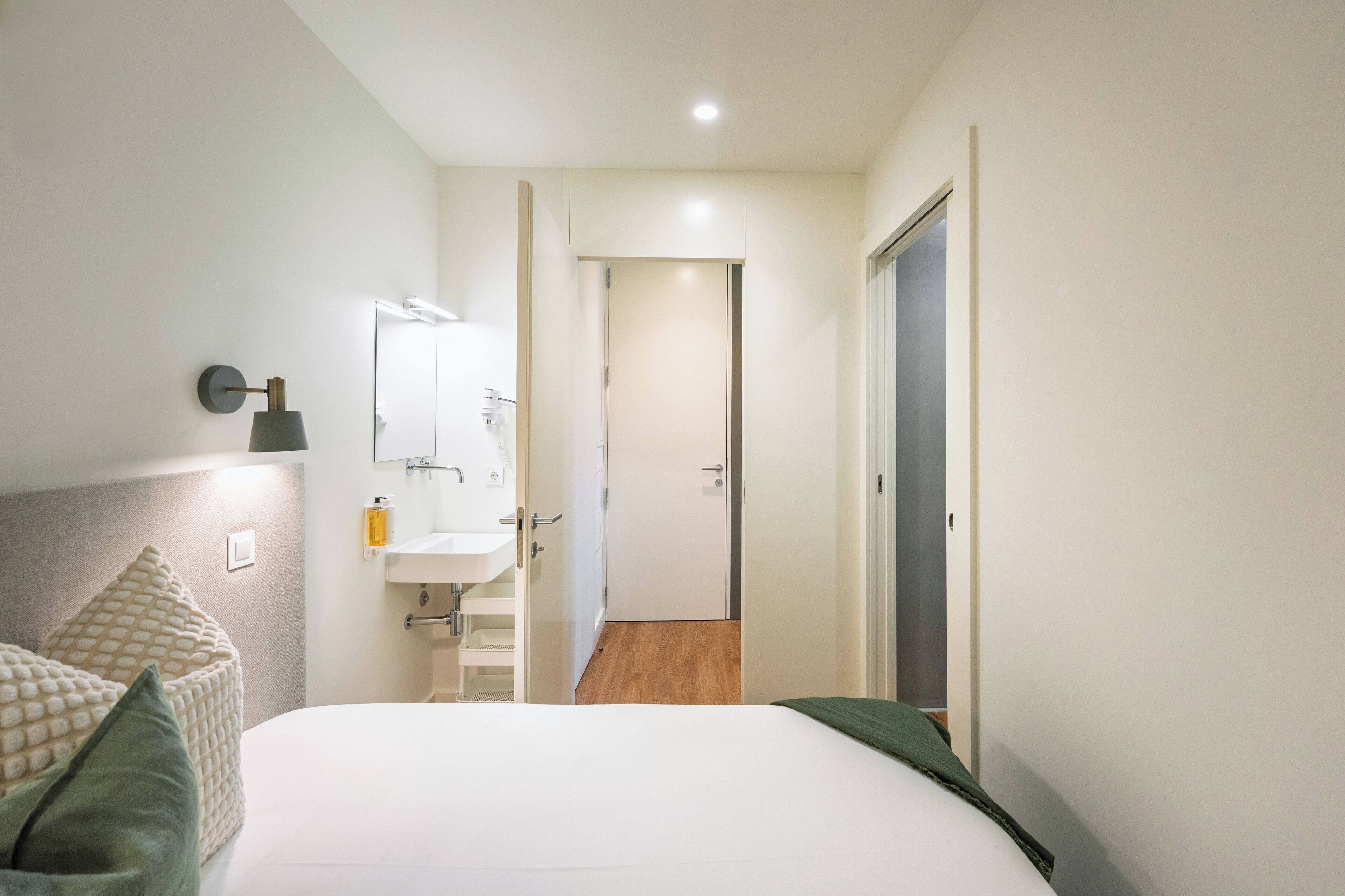 A hotel room with a bed, a bedside table, a mirror, a wall-mounted light, and a door leading to another room.