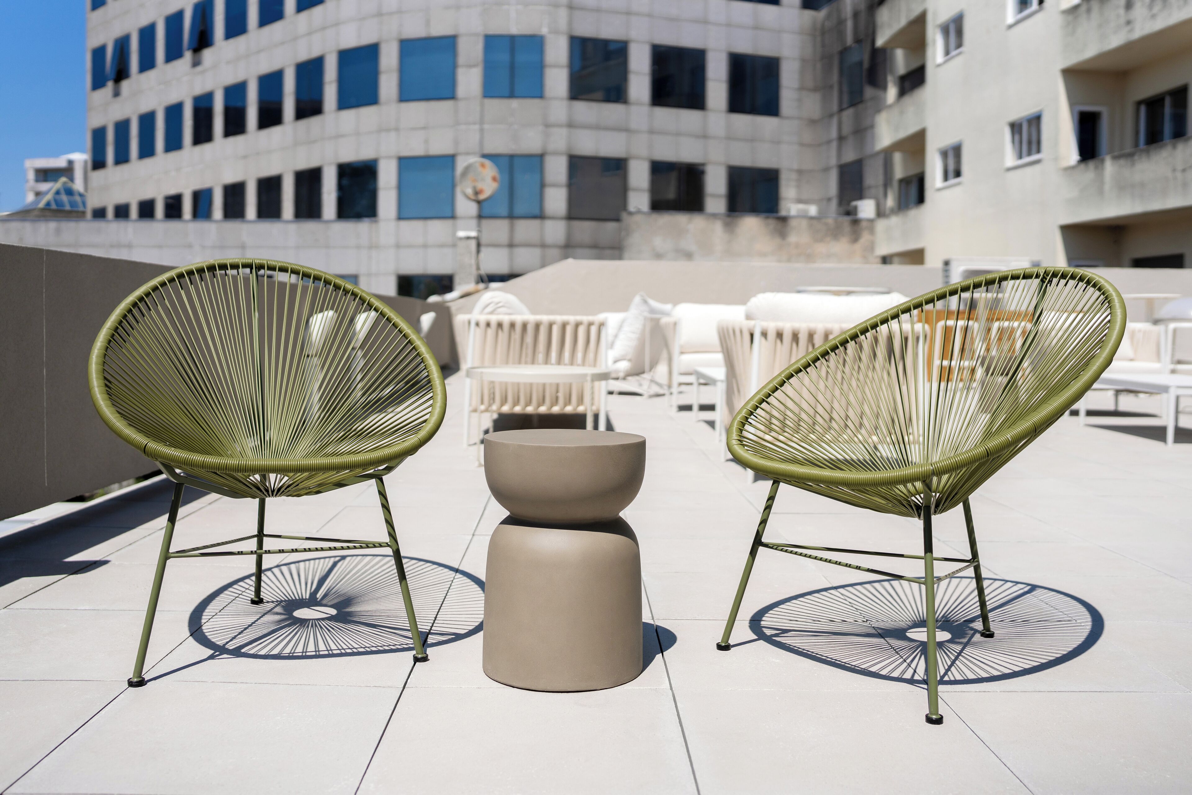 A rooftop terrace with two green wire-framed chairs, a beige table, and white lounge chairs.